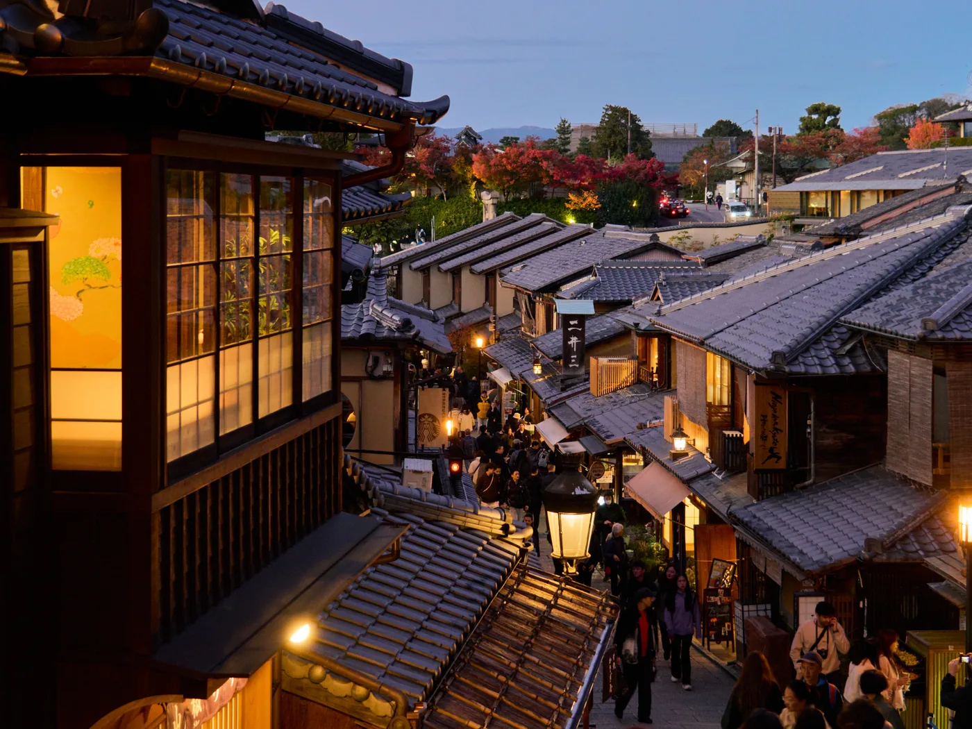 Visitors on the steps of Ninenzaka in Kyoto, surrounded by traditional tiled roofs and warm evening lights.