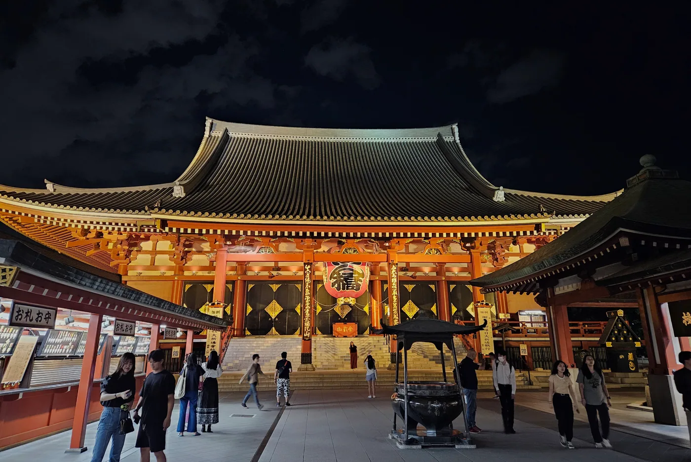 The illuminated main hall of Senso-ji in Asakusa with visitors in the foreground at night.